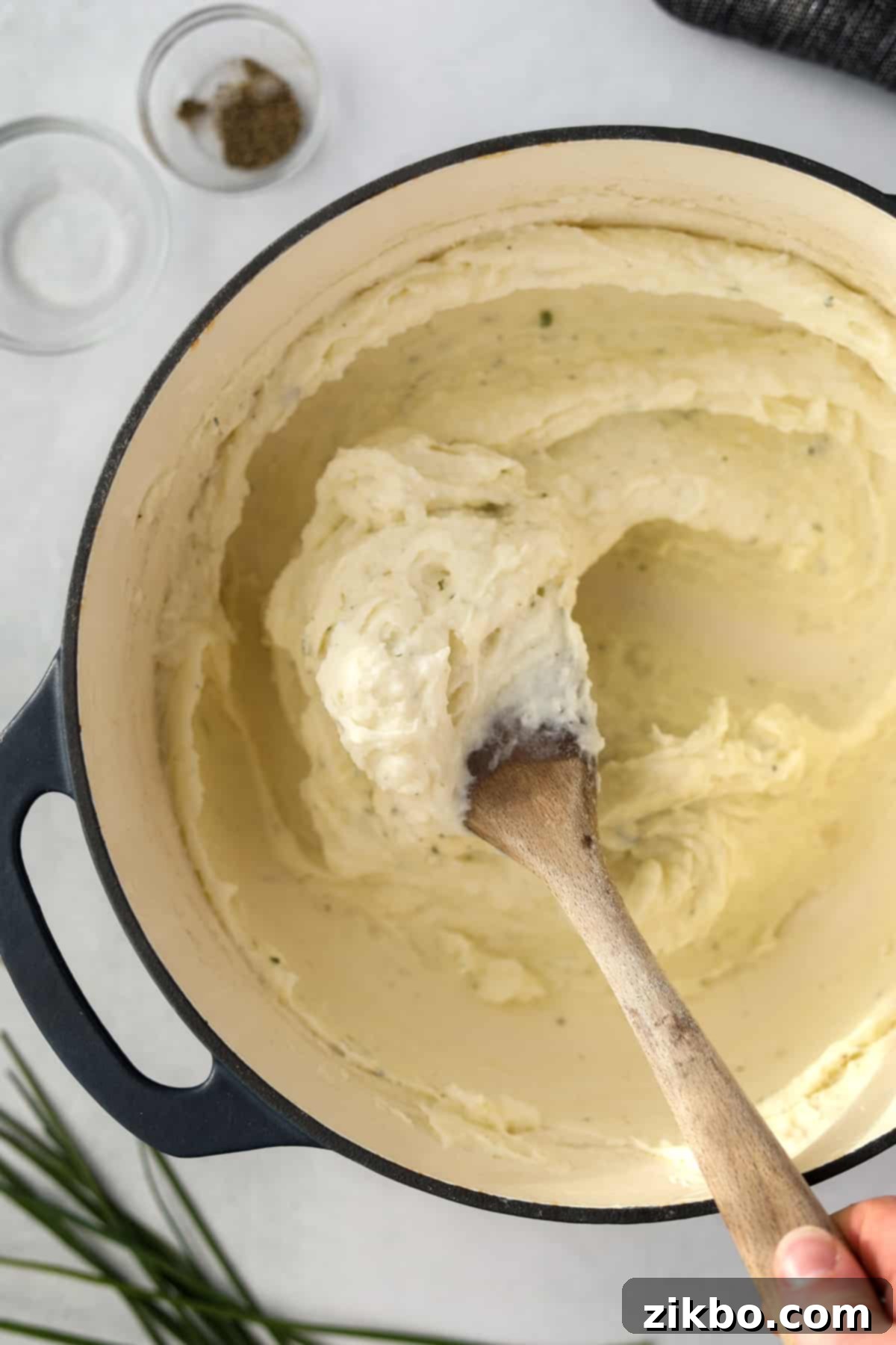 A wooden spoon actively whipping and stirring mashed potatoes in a Dutch oven, incorporating the melted Boursin cheese.