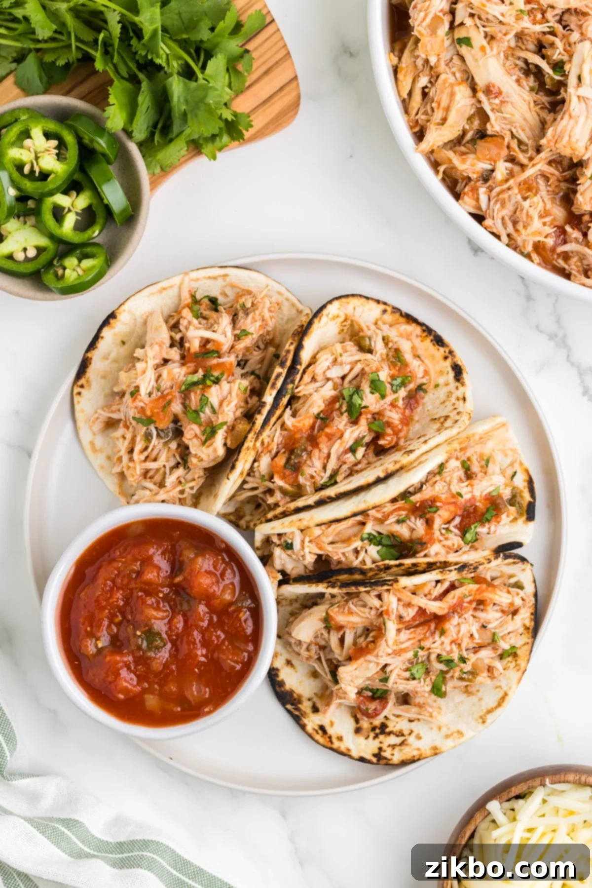 A plate filled with four chicken tacos, topped with cilantro and a squeeze of lime, with a bowl of salsa on the side.