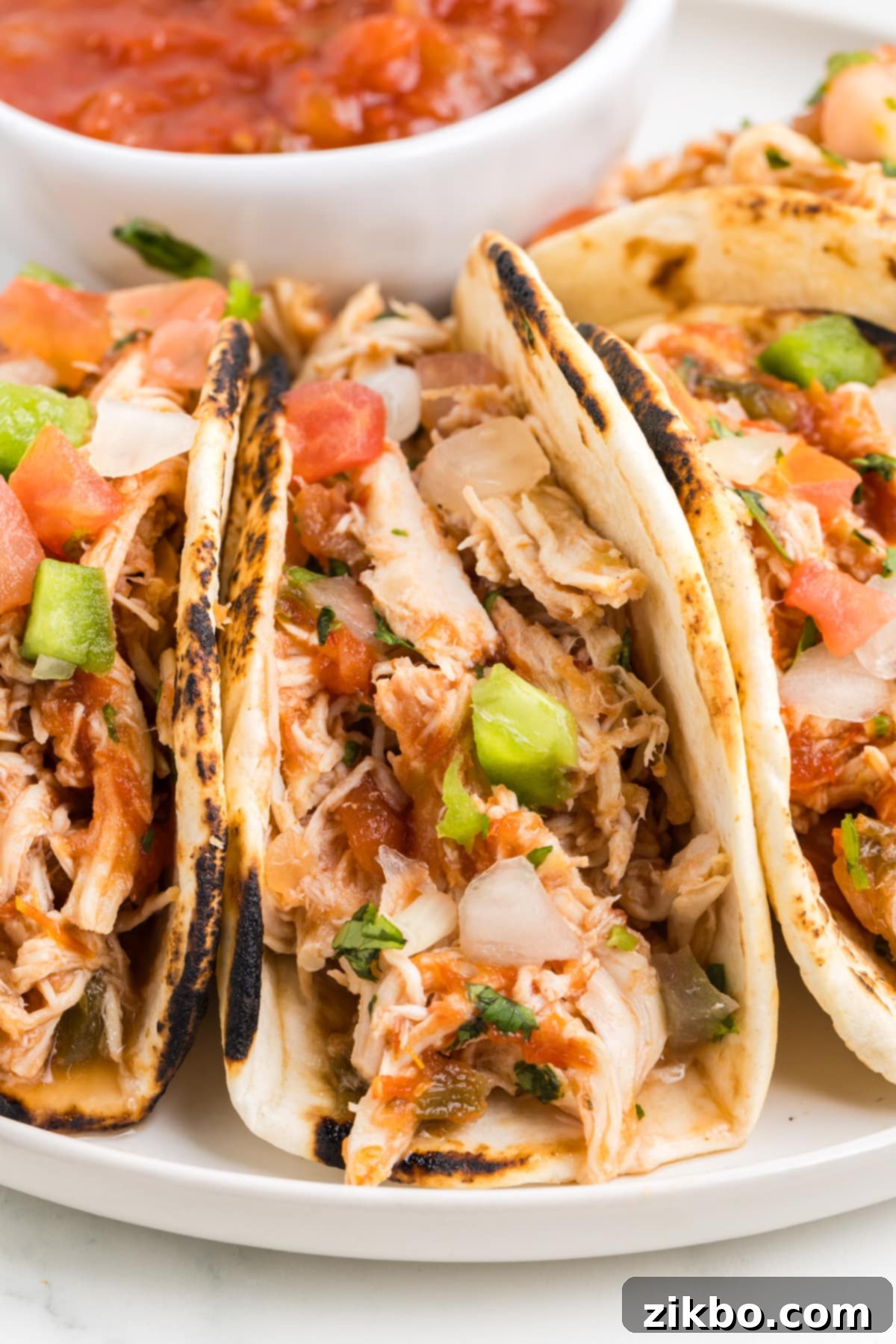 A close-up shot of a plate of small, enticing chicken tacos, beautifully topped with fresh cilantro and finely chopped tomato, highlighting their delicious texture.