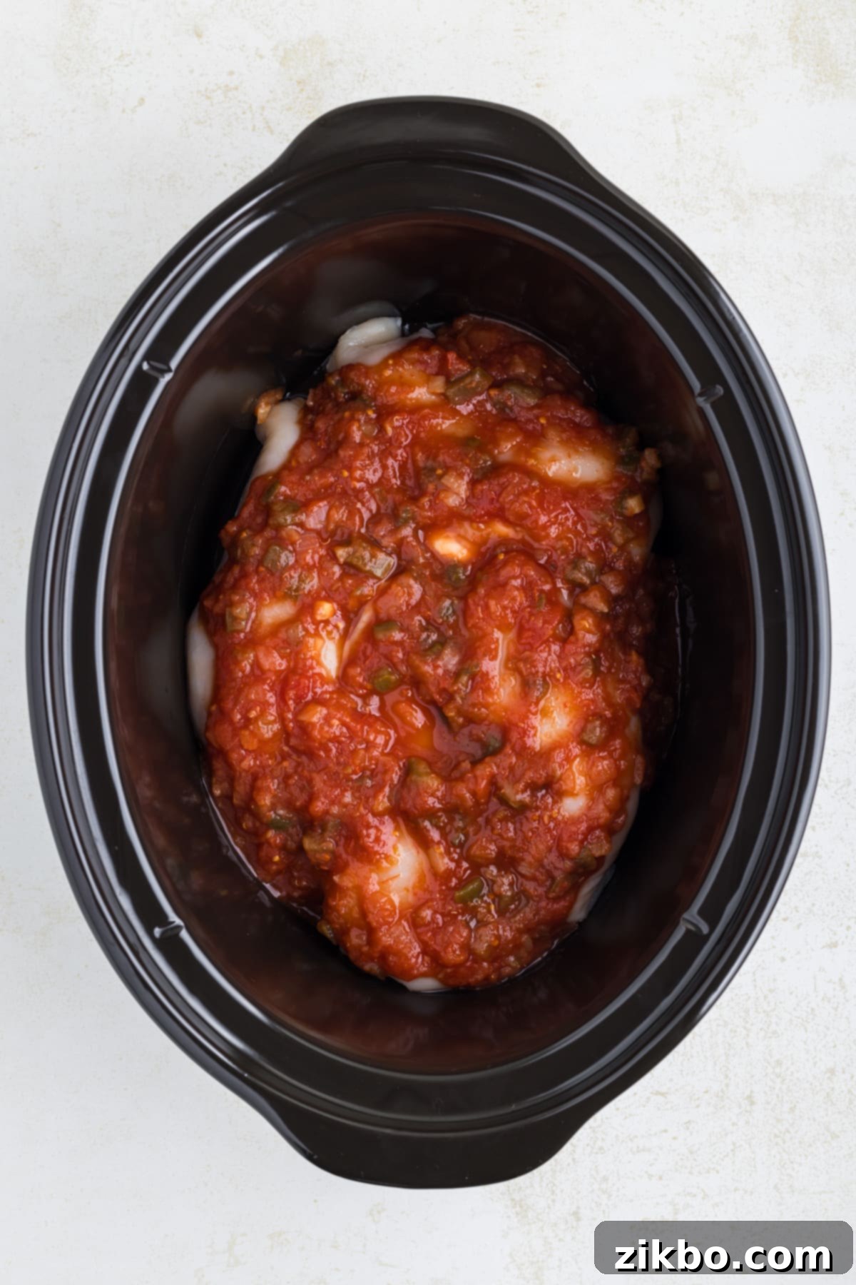 A slow cooker filled with boneless, skinless chicken breasts and generously covered with salsa, ready for cooking.
