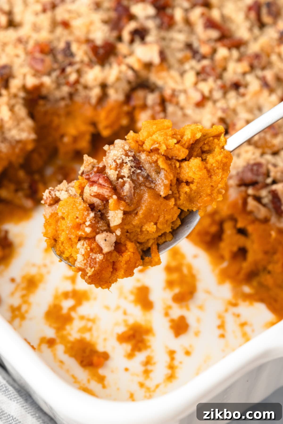 A spoon taking a portion of sweet potato casserole with pecans, showcasing the golden-brown topping and soft interior.