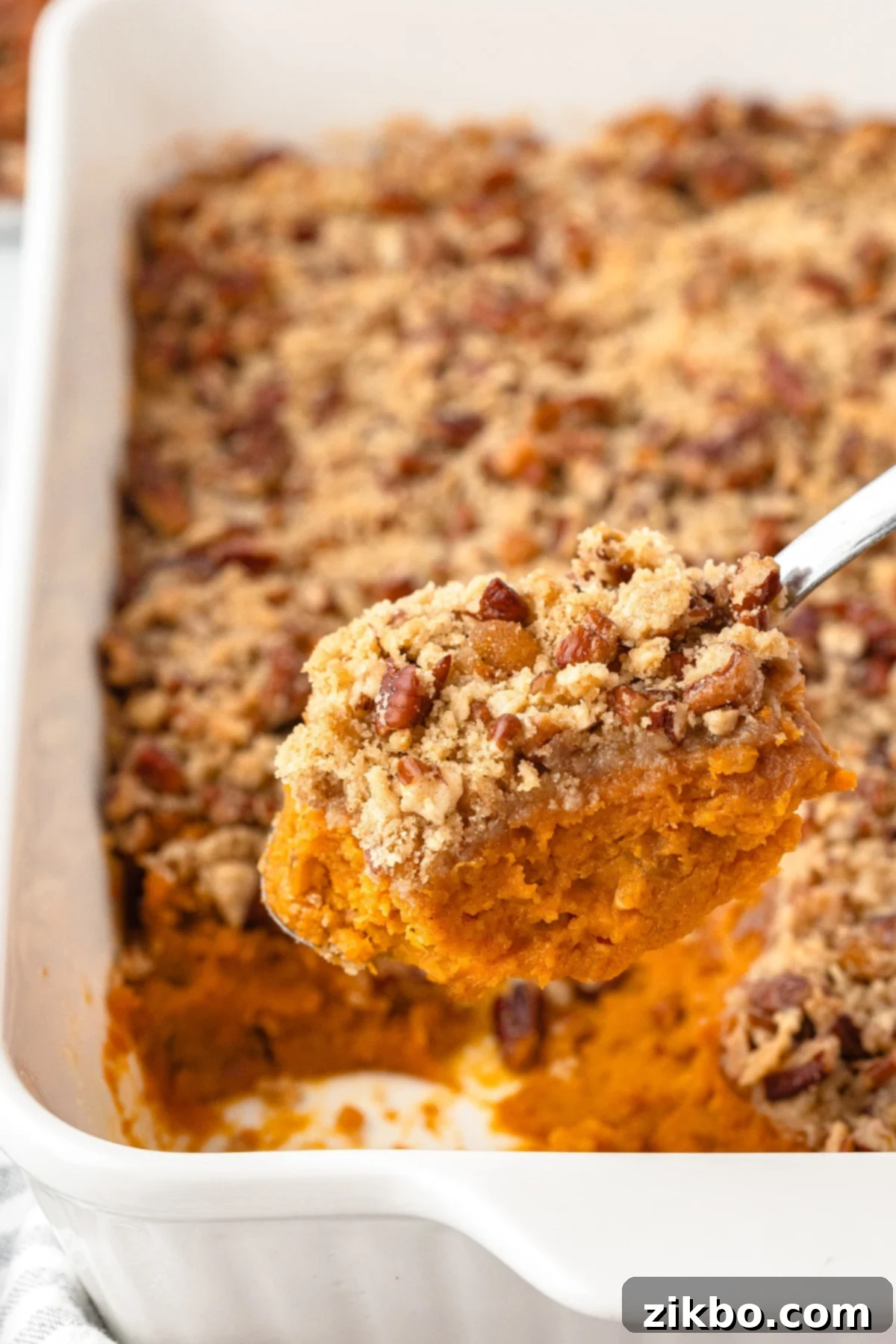 A spoon scooping a portion of sweet potato casserole from a baking dish, highlighting the texture of the filling and topping.