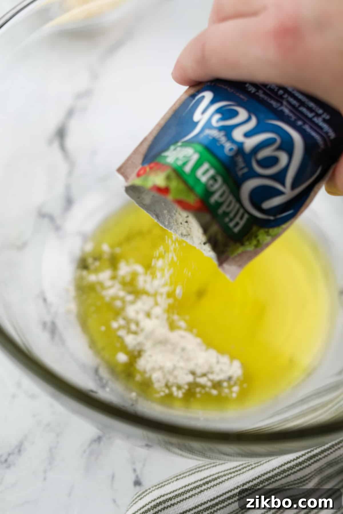 Sprinkling a packet of dry ranch seasoning mix into olive oil in a large bowl.