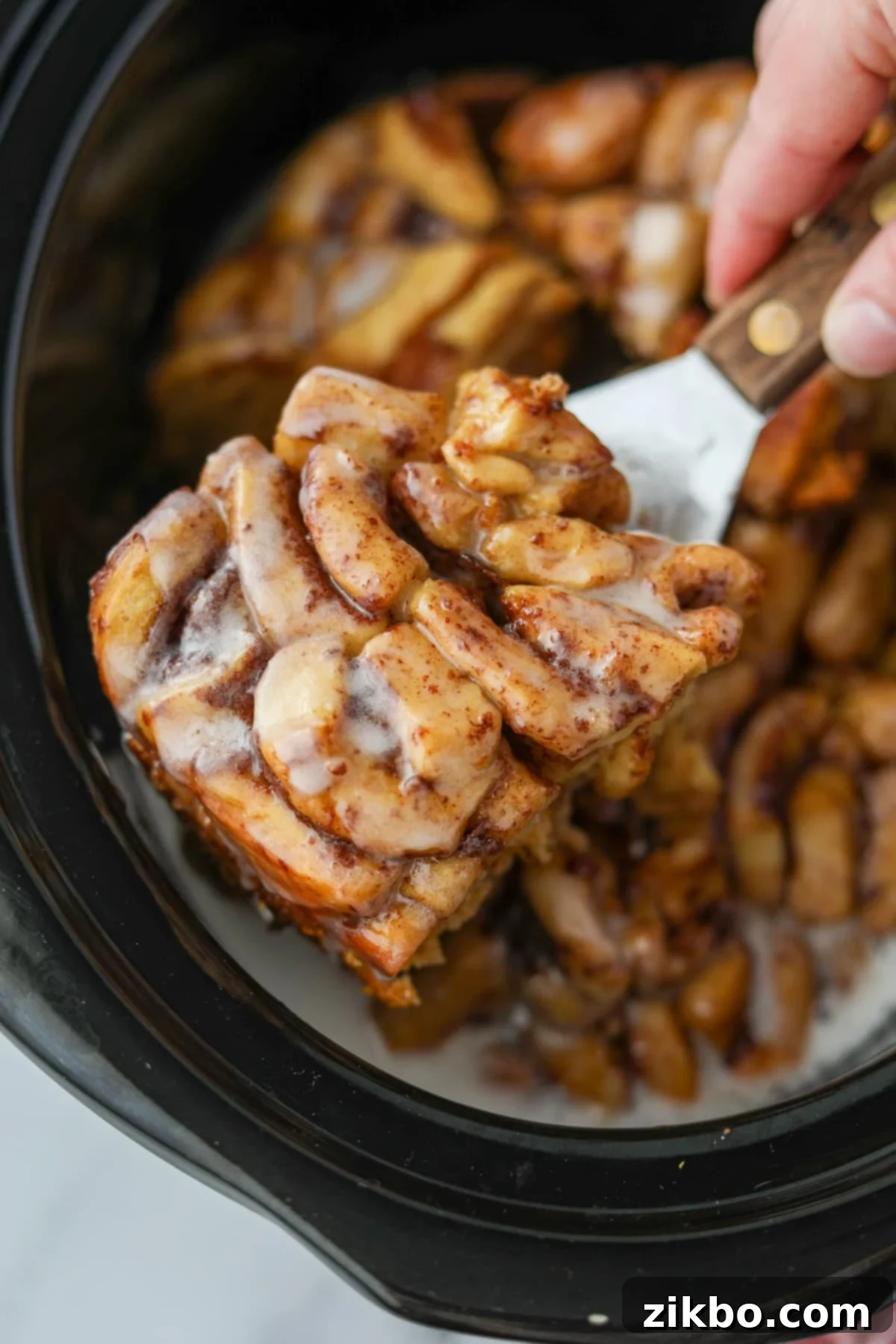 Taking a portion of crock pot cinnamon rolls from a slow cooker. Golden brown cinnamon roll casserole with icing drizzle.
