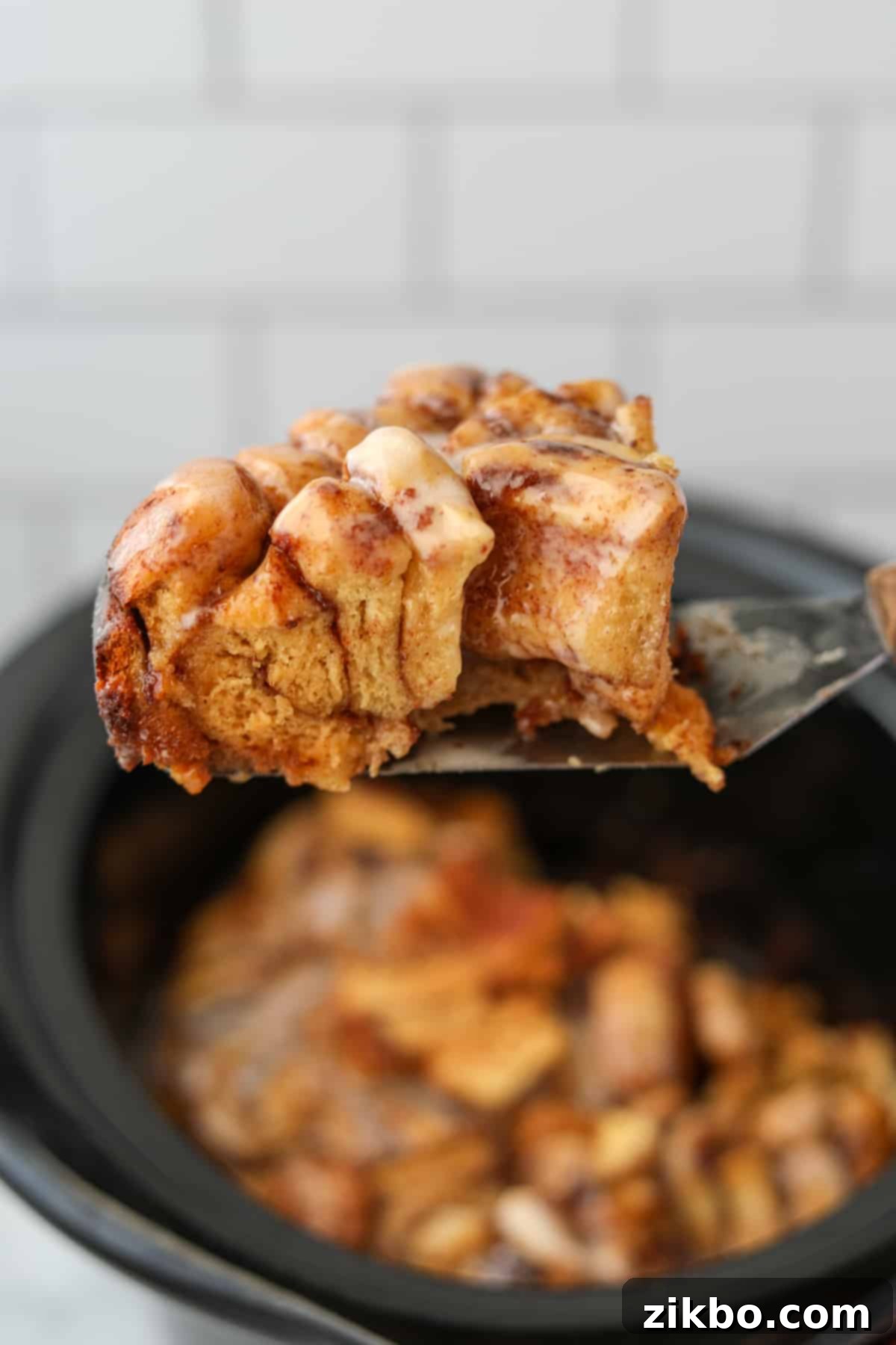 Lifting a slice of cinnamon roll casserole from a crock pot, topped with generous icing.