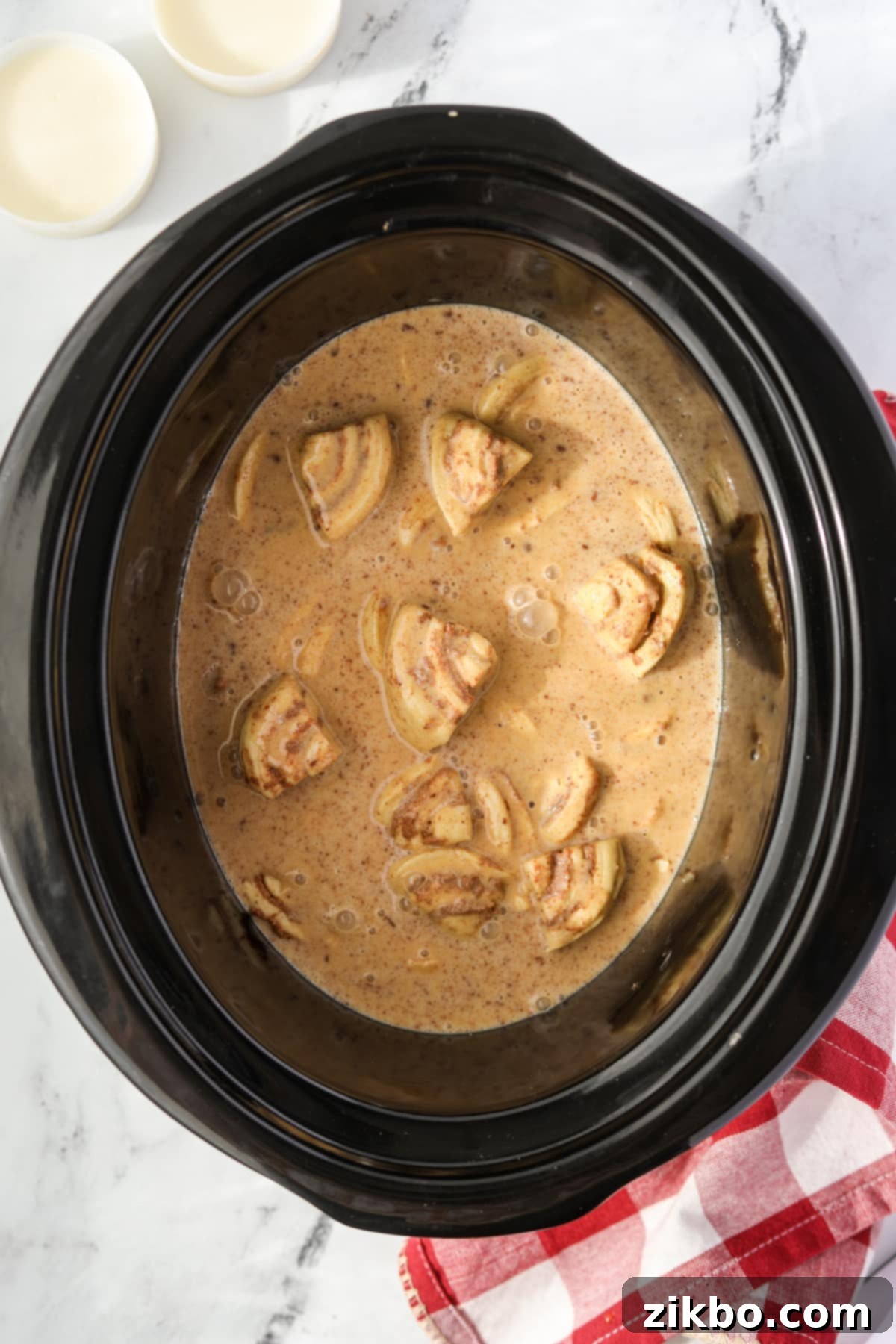 A slow cooker filled with cinnamon roll pieces and a custard mixture poured over them.