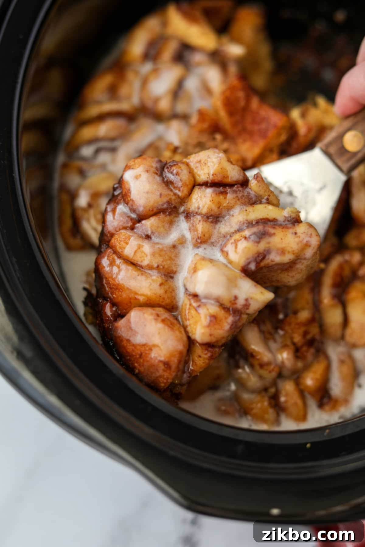 Lifting a slice of cinnamon roll bake from a crock, showing the gooey, cooked texture.