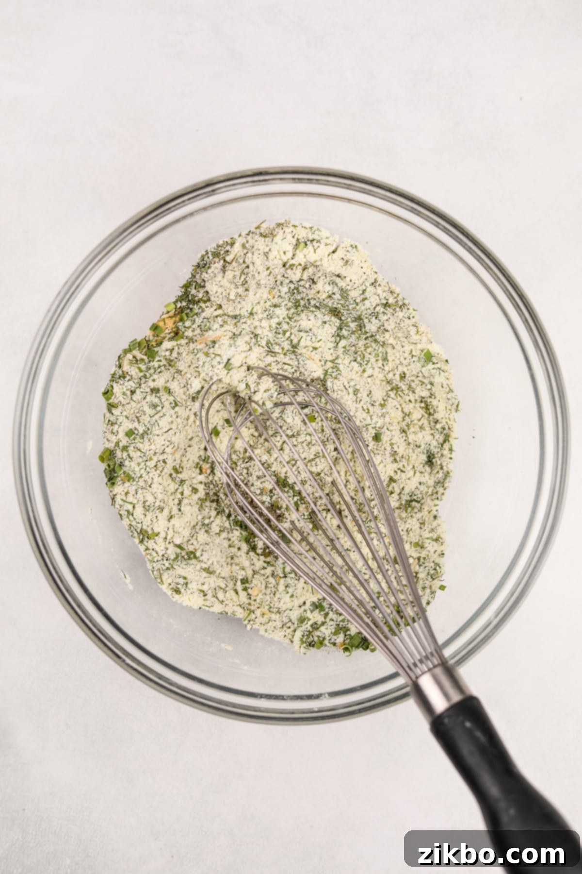 A hand whisking together dried herbs and buttermilk powder in a bowl, creating the homemade ranch seasoning mix.