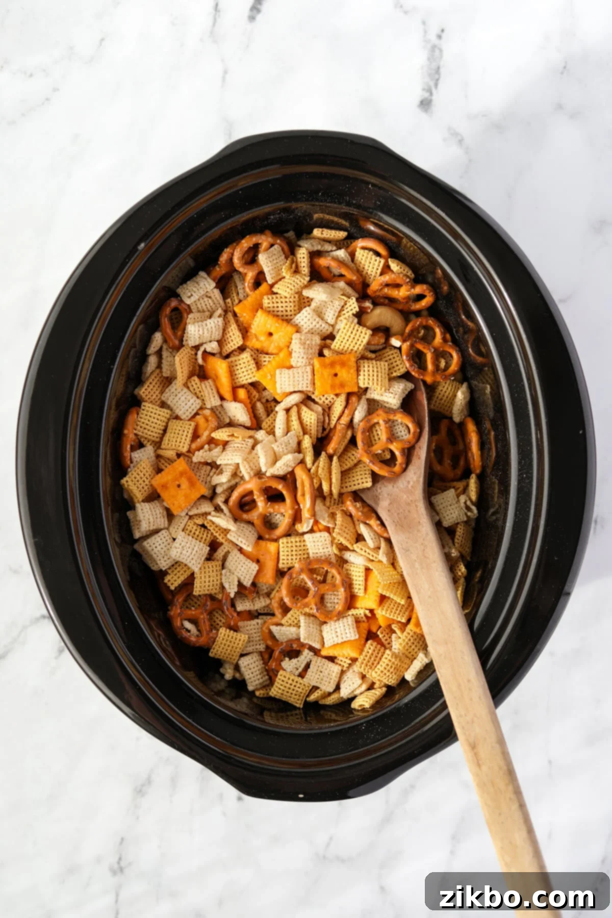 A wooden spoon gently stirring Chex cereal, pretzels, and nuts in a slow cooker to combine them evenly.