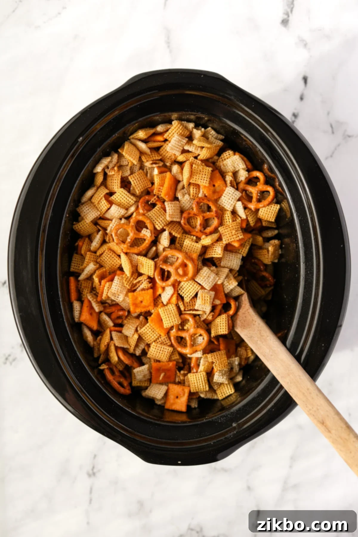 A wooden spoon stirring fully coated Chex Mix ingredients in a slow cooker before cooking.
