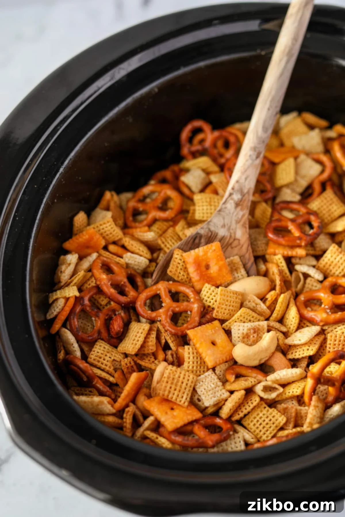 A wooden spoon stirring the finished, golden brown Chex Mix in a slow cooker after it has cooked, showing its crispy texture.