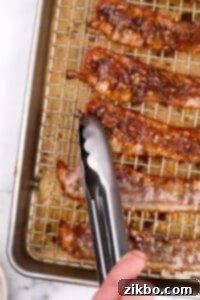A pair of tongs flipping bacon on a baking sheet, showing the caramelized side and the side to be coated.