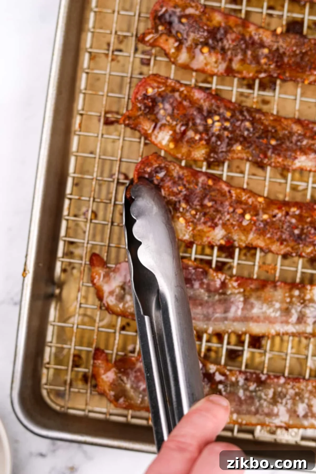 A pair of tongs flipping bacon on a baking sheet, showing the caramelized side.