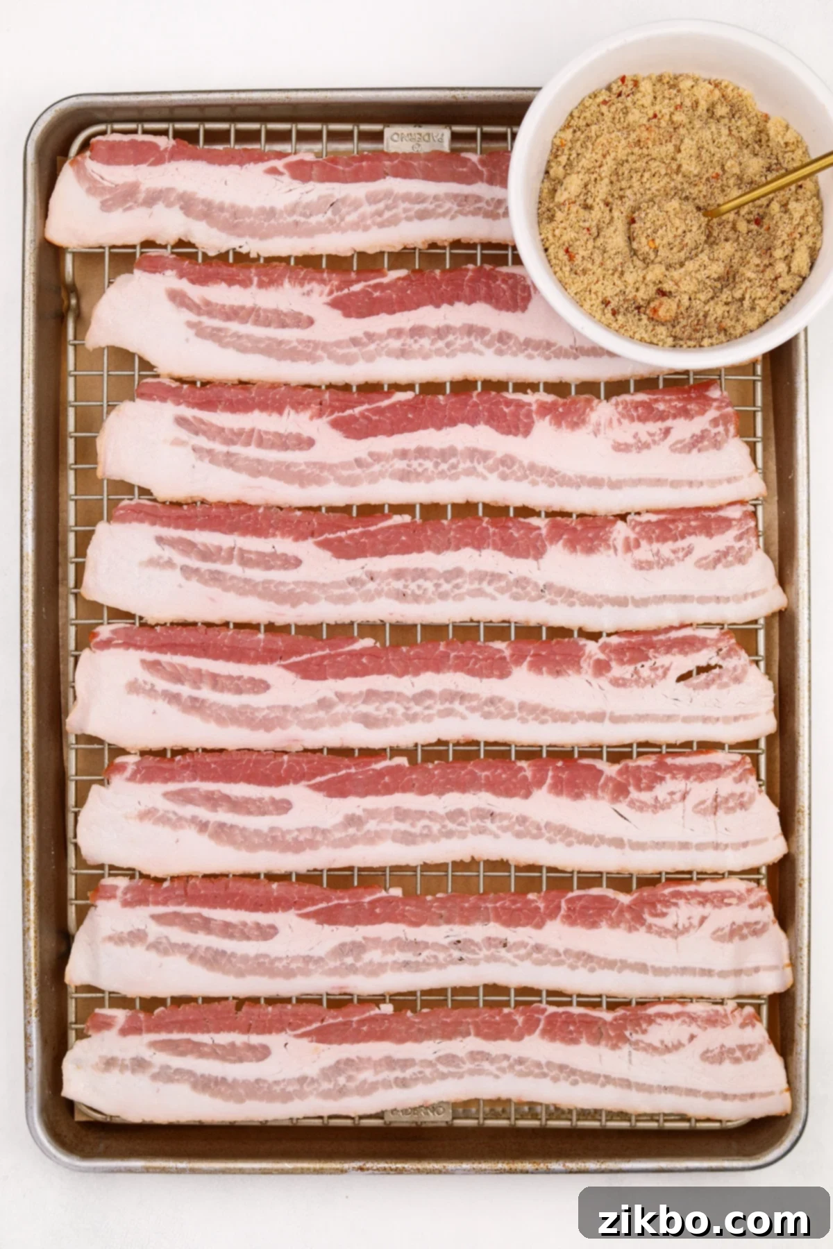 A baking sheet with slices of bacon neatly spread out on a wire rack, with a small bowl of brown sugar spice mix beside it.