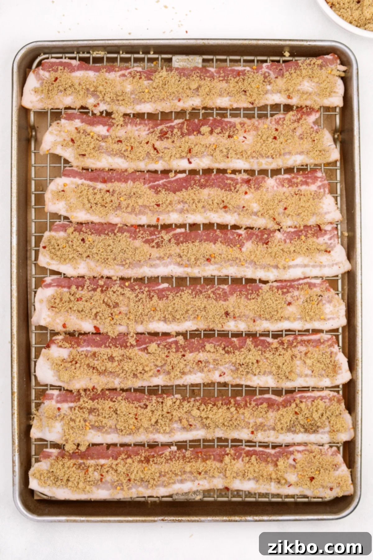 A baking sheet with bacon slices on a wire rack, generously topped with a brown sugar spice rub.