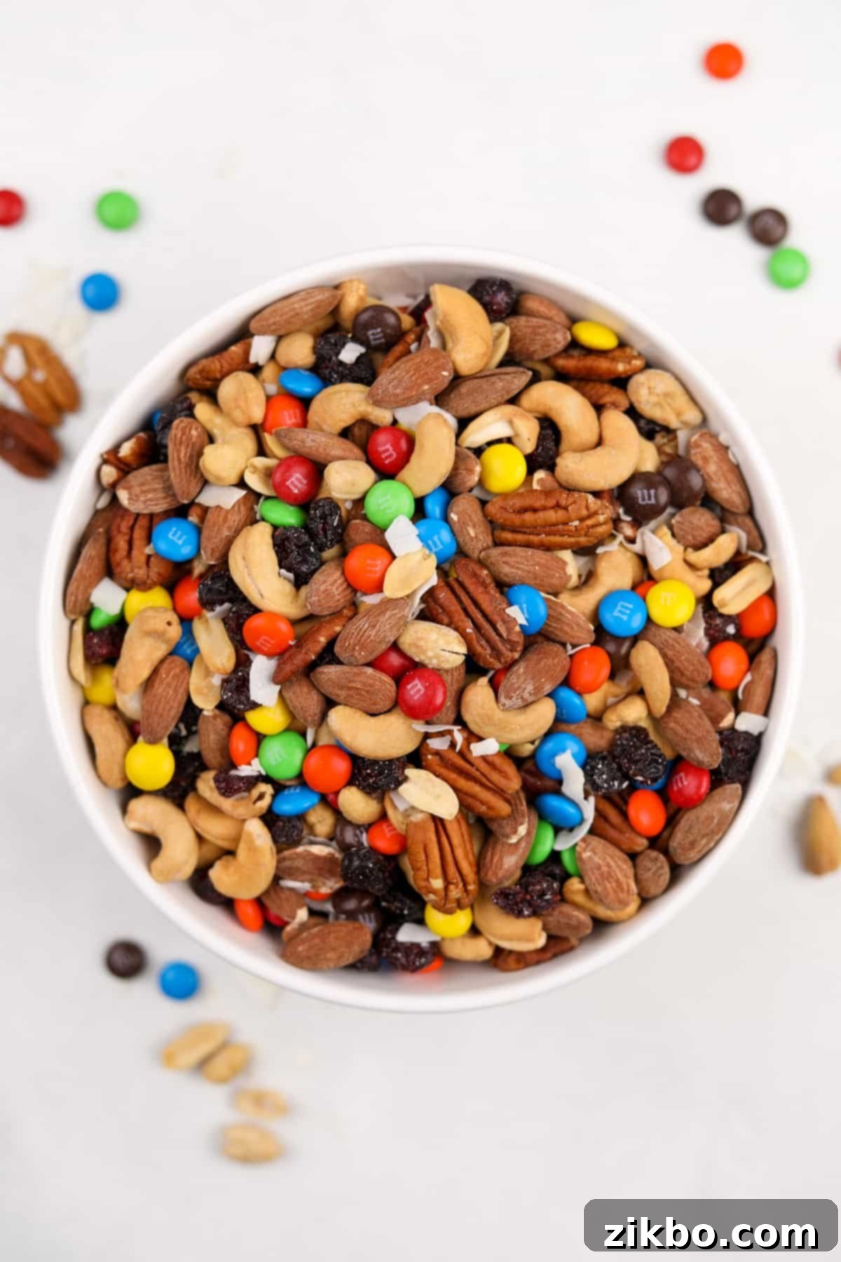 A large bowl of vibrant homemade trail mix on a white surface, showcasing a colorful assortment of nuts, dried fruits, and M&M's.