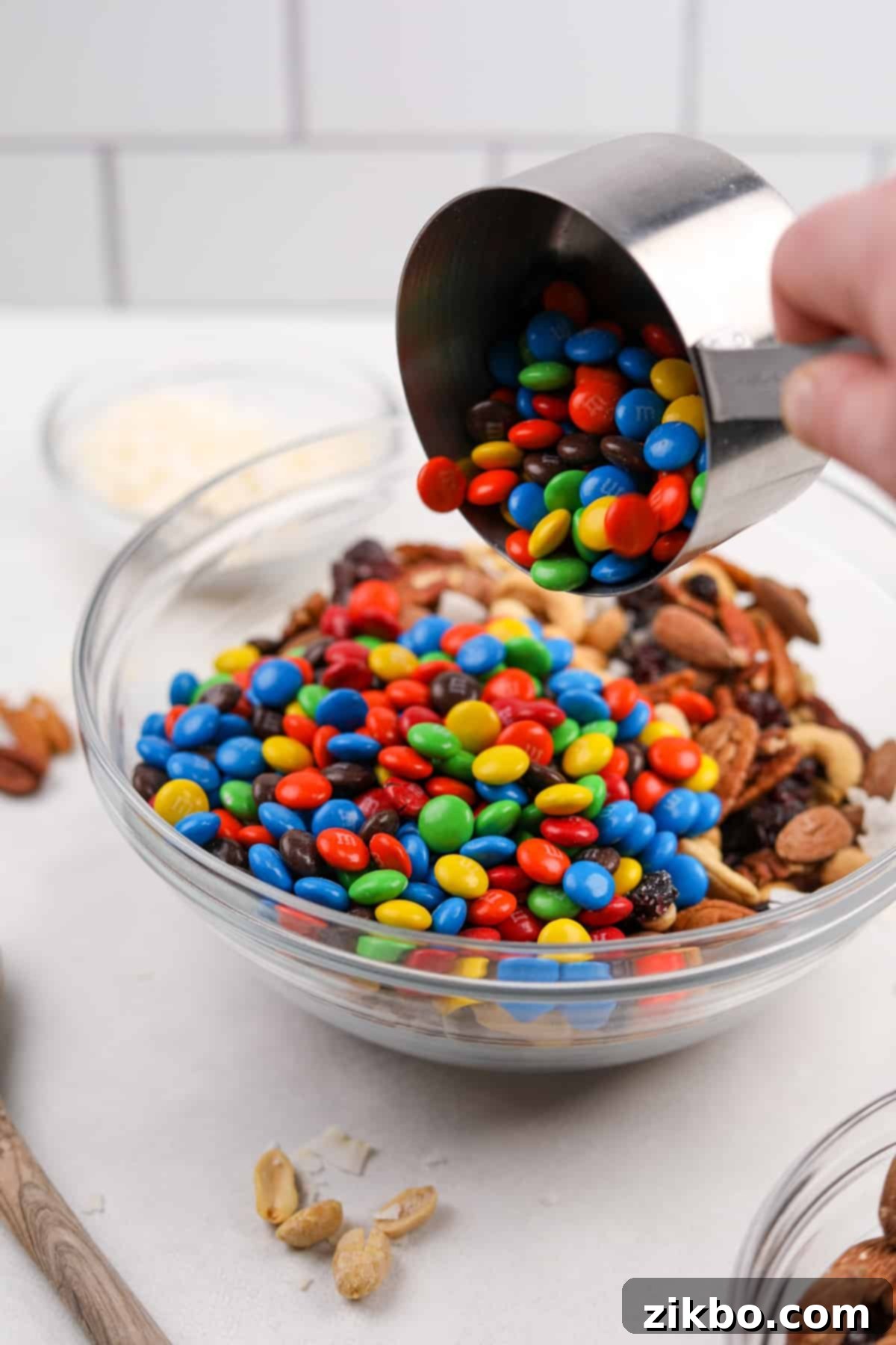 Person pouring colorful M&M's into a bowl of mixed nuts and dried fruit for trail mix.