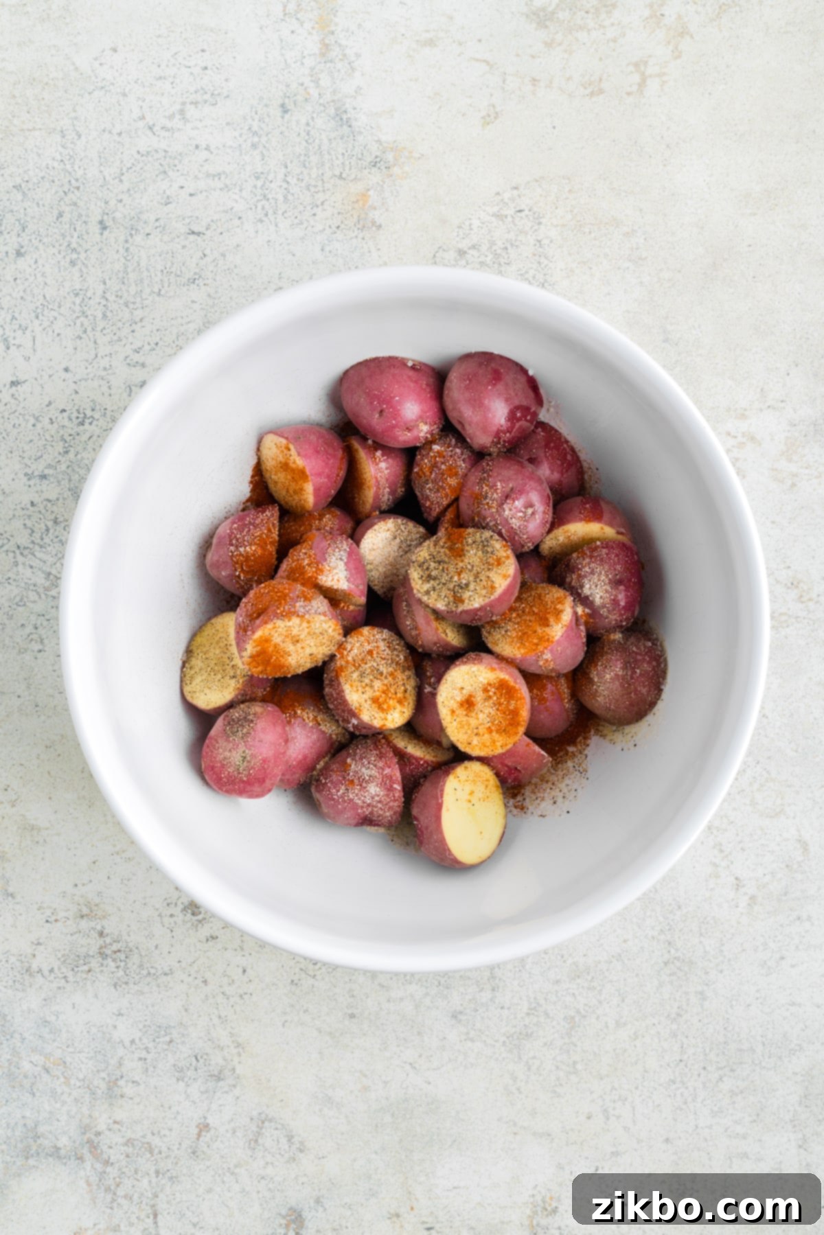 A bowl of cut red potatoes generously topped with olive oil and a vibrant blend of seasonings.